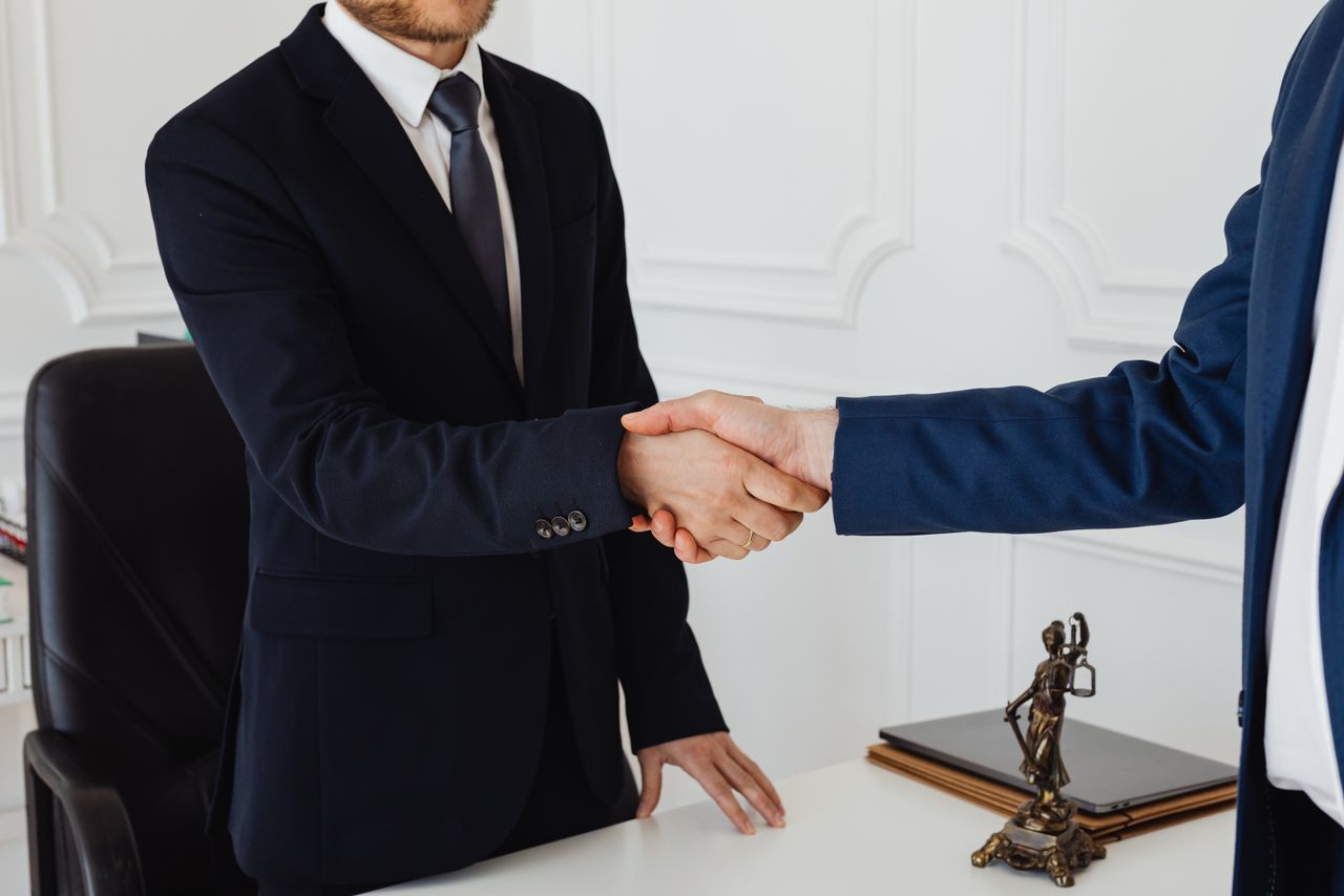 about-us Two men in suits shaking hands in a formal office environment symbolizing an agreement.
